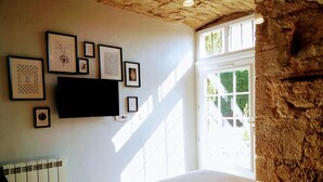 A bedroom with a bed, a TV mounted on the wall, and several framed pictures. There's a window with a view of greenery outside. - La Meridienne des Collines (La Motte-de-Galaure)
