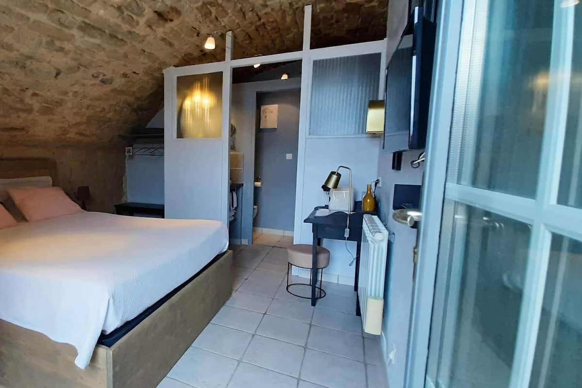 A bedroom with a stone ceiling, a bed with white bedding, a small table with a lamp, and a sliding glass door leading to a bathroom.
