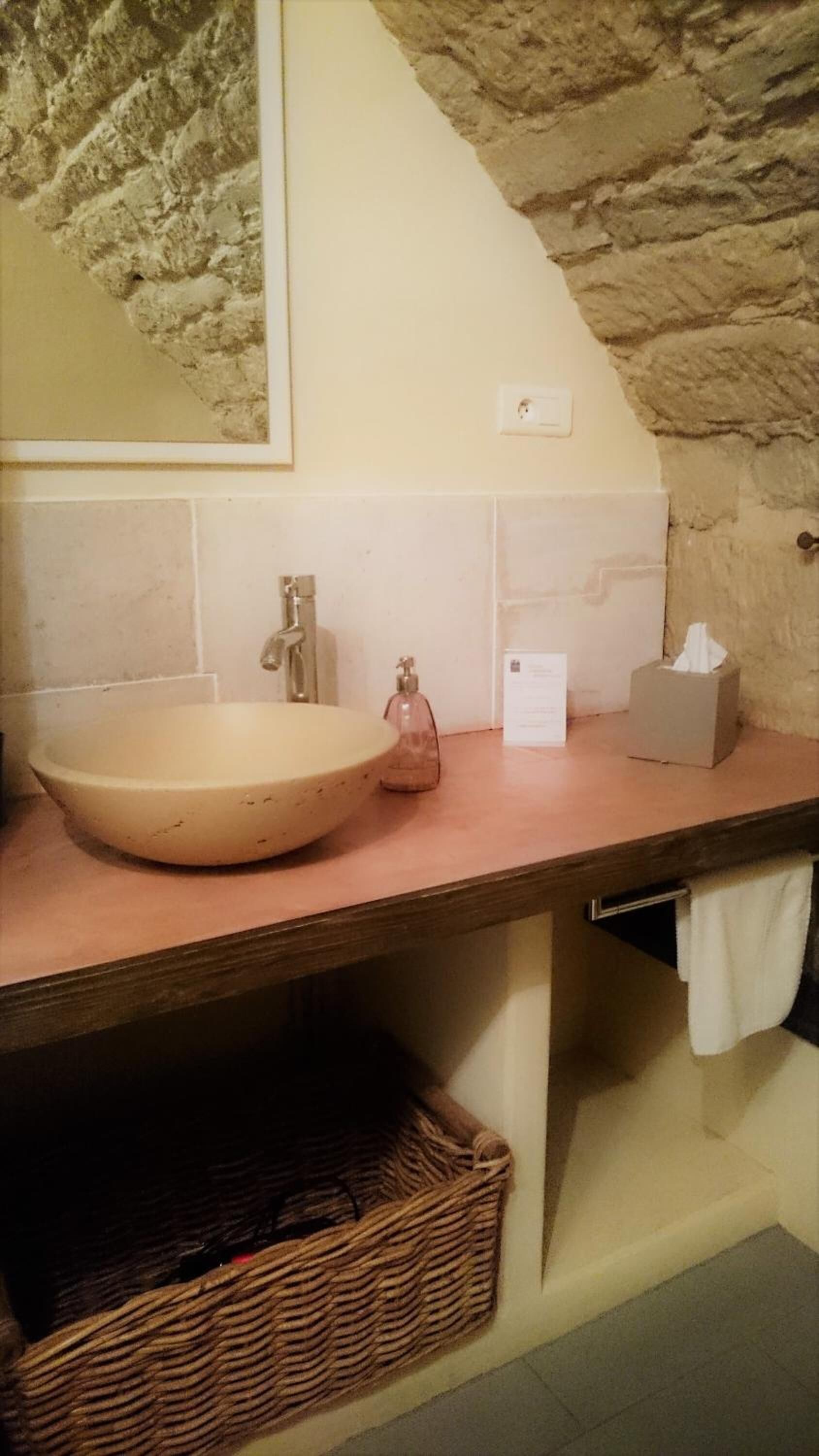 A bathroom with a stone wall, a round sink, a mirror, a towel rack, and a wicker basket.