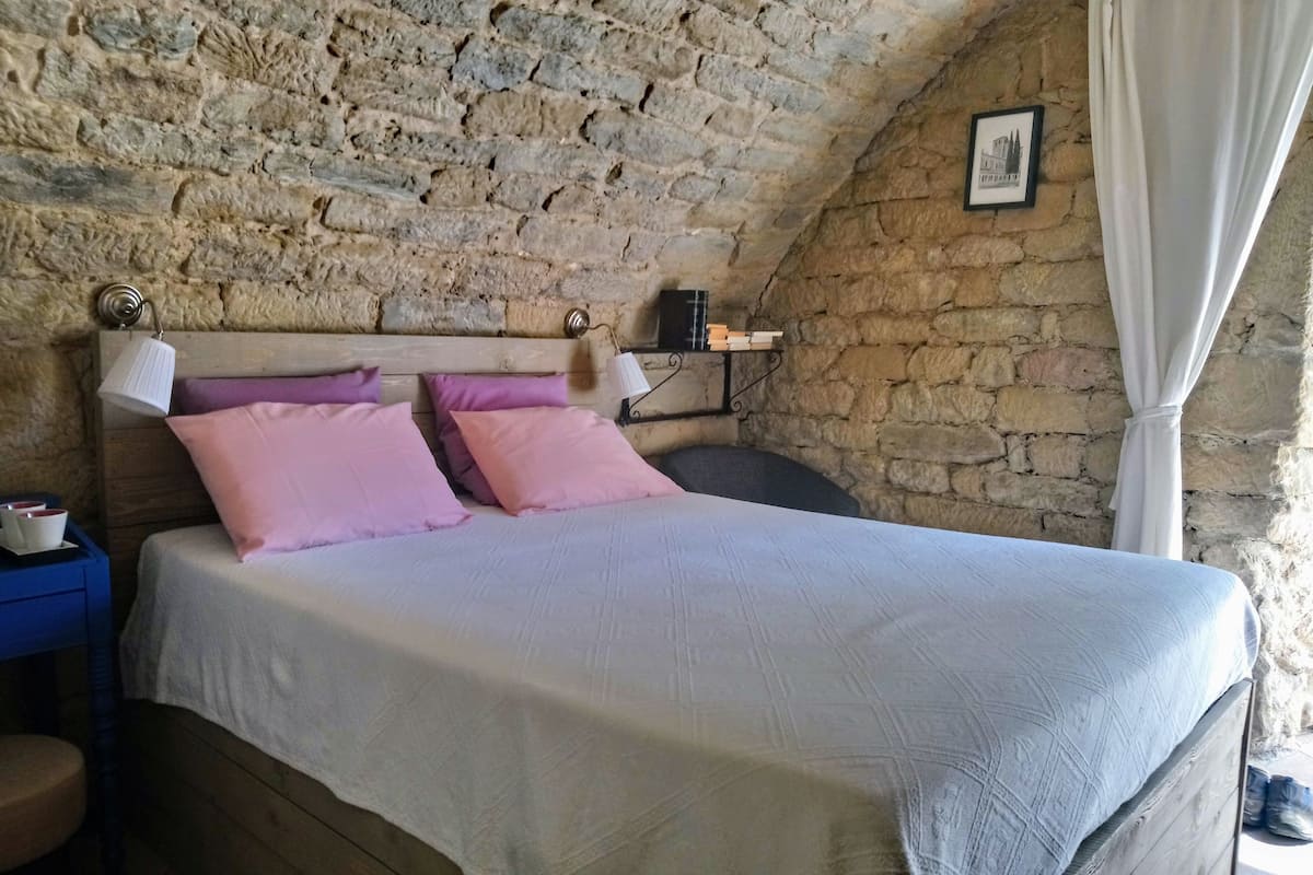 A bedroom with a stone wall, a wooden headboard, a bed with white and pink bedding, a nightstand with a lamp, and a small shelf with books.