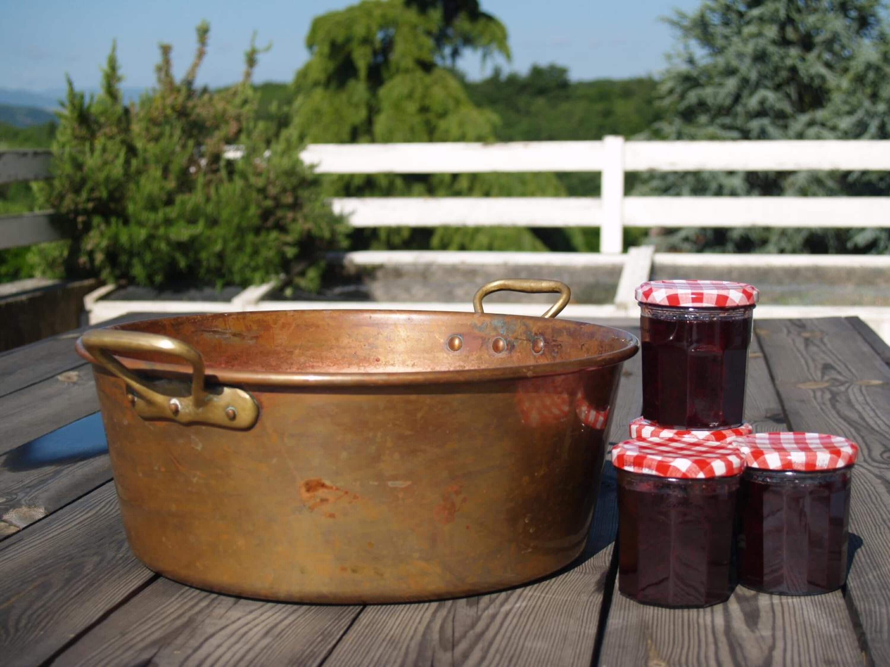 Un décor rustique en plein air, avec un grand pot en cuivre, deux bocaux en verre dotés de couvercles à carreaux rouges et blancs, et une clôture blanche en arrière-plan.