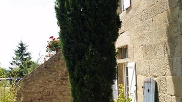 Un edificio de piedra con un árbol alto y esbelto en primer plano. Hay plantas en macetas y una pequeña mesa con sillas afuera.