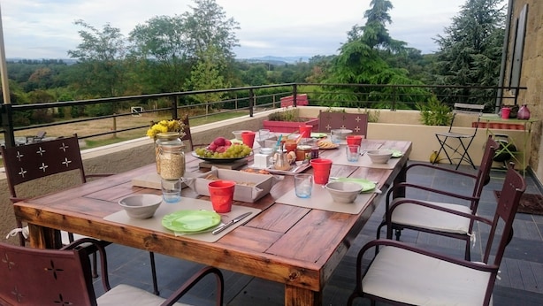 An outdoor dining area with a wooden table set for a meal, surrounded by chairs, and overlooking a scenic landscape. - La Meridienne des Collines (La Motte-de-Galaure)