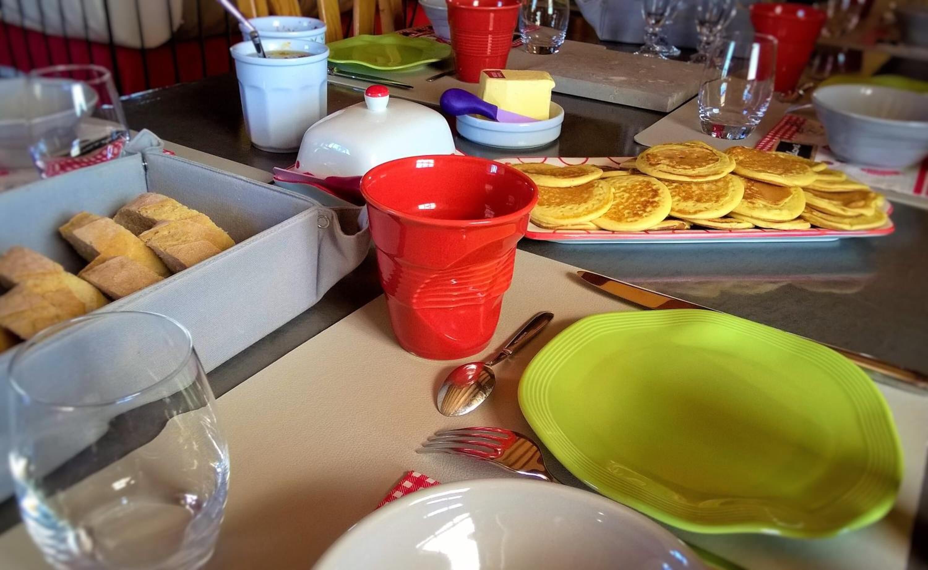 Une table de petit-déjeuner avec des crêpes, une tasse rouge, une assiette verte et une boîte de biscuits.