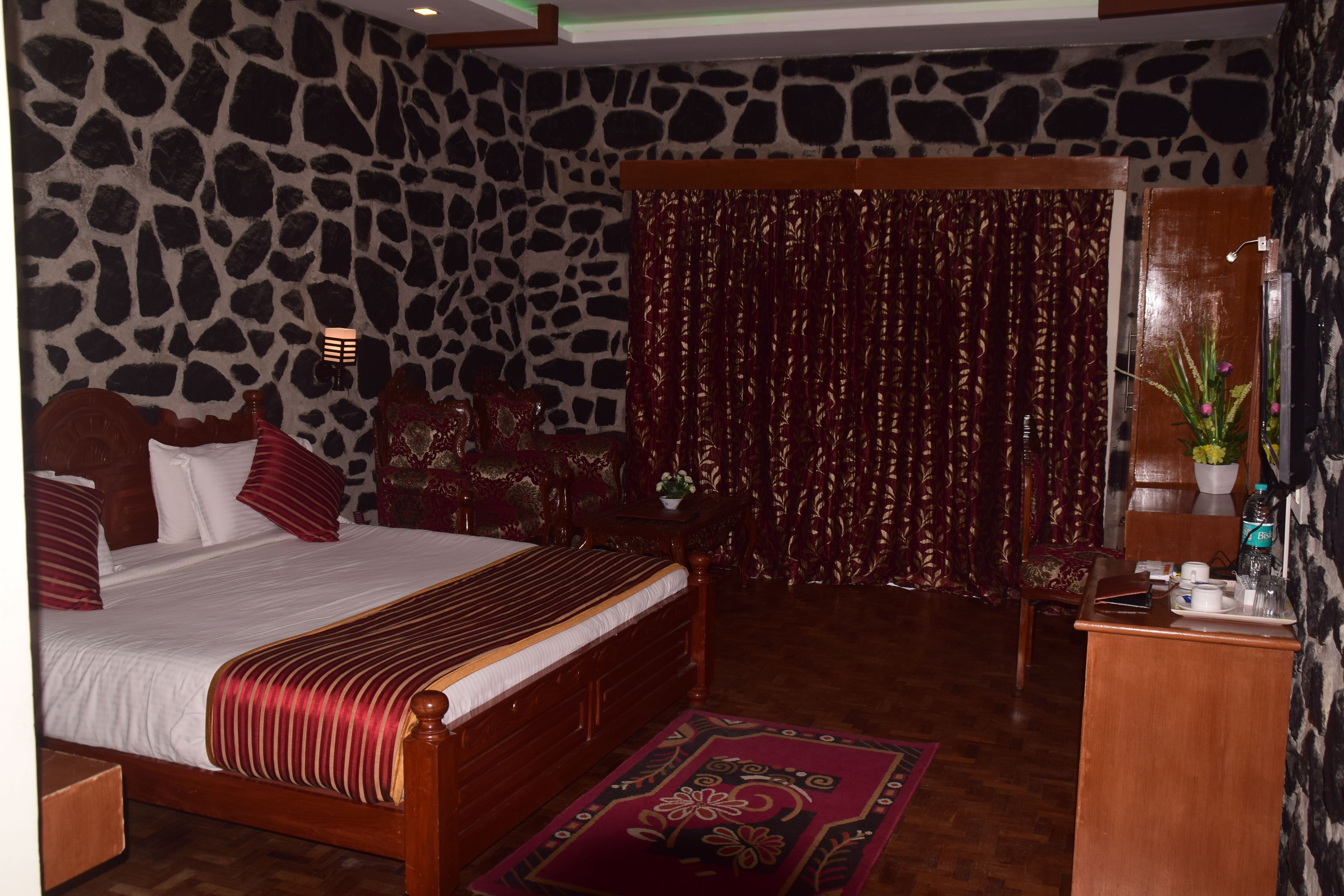 A bedroom with a stone-patterned wall, a bed with red and white bedding, a wooden headboard, a red curtain, a small table with a plant, and a desk with a water bottle.