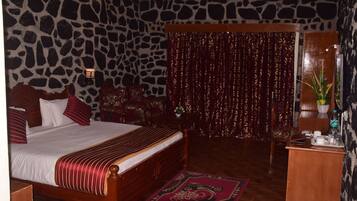 A bedroom with a stone-patterned wall, a bed with red and white bedding, a wooden headboard, a red curtain, a small table with a plant, and a desk with a water bottle.