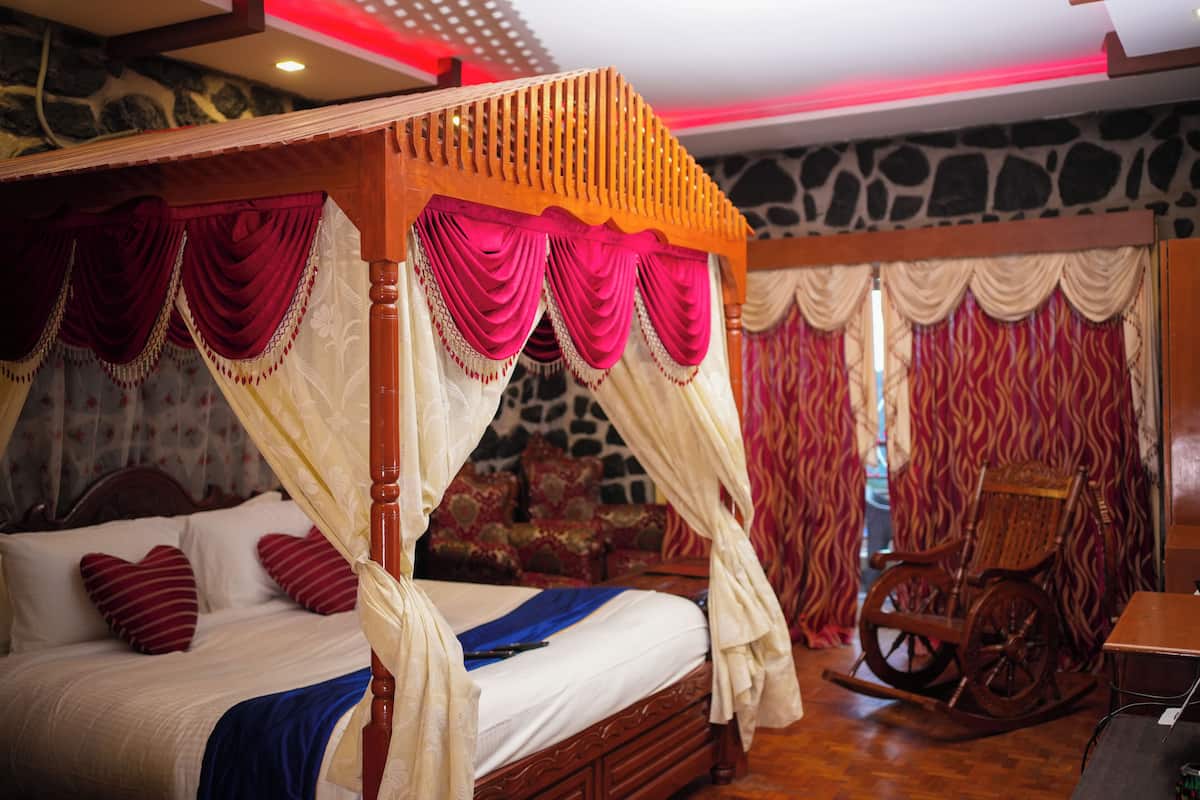 A four-poster canopy bed with red and white drapery, a rocking chair, and a stone wall backdrop.