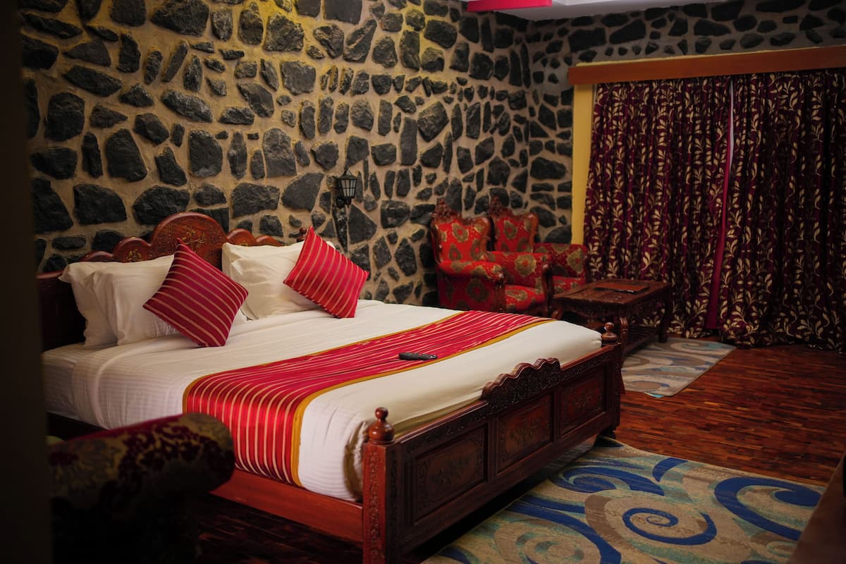 A bed with a wooden frame and red bedding, a stone wall, a patterned curtain, and a small table with a lamp.