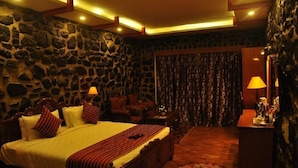 A bedroom with a stone wall, a bed with a striped bedspread, a wooden headboard, two bedside lamps, a patterned curtain, and a small table with a lamp. - Jem Valley (Kodaikanal)