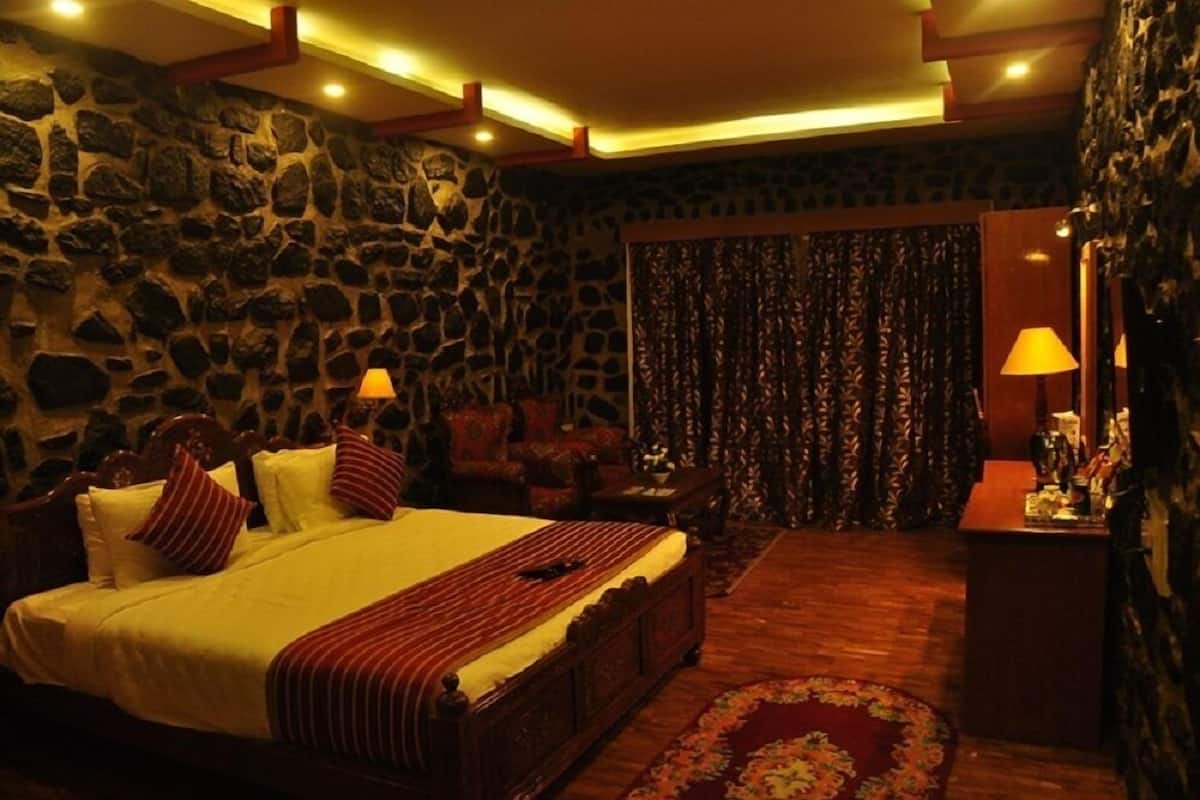 A bedroom with a stone wall, a bed with a striped bedspread, a wooden headboard, two bedside lamps, a patterned curtain, and a small table with a lamp.