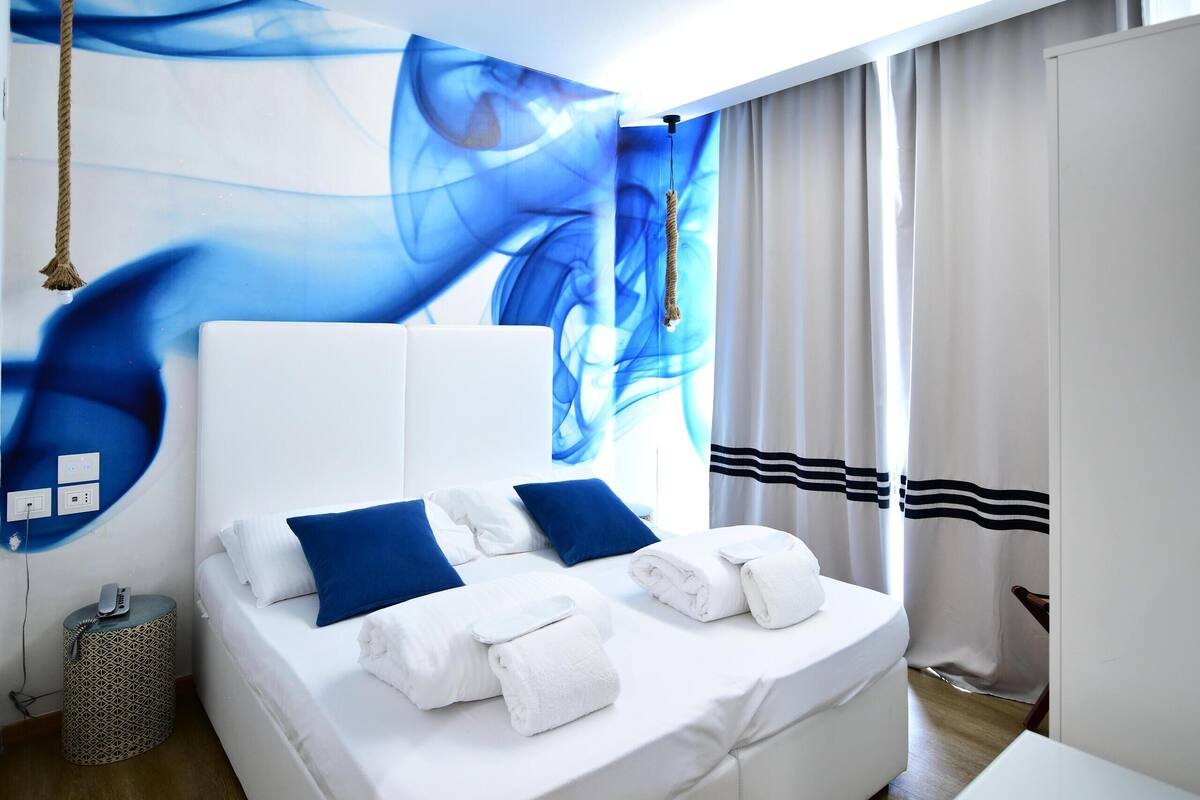 Una habitación de hotel moderna con una cama blanca, almohadas azules y un amplio mural abstracto en la pared.