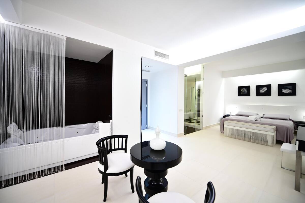 Una habitación de hotel moderna con una bañera grande, una zona de comedor con una mesa y sillas negras, y una cama con cabecero.