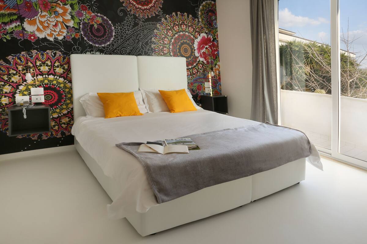 Un dormitorio moderno con una cama grande, un tapiz colorido en la pared y vistas al exterior.