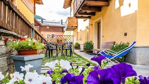 A courtyard with a wooden table and chairs, a hammock, and potted plants with flowers. - Chalet Mottolino (Livigno)