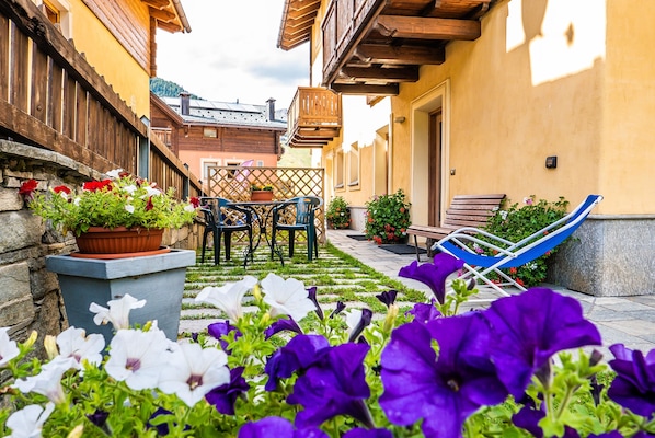 A courtyard with a wooden table and chairs, a hammock, and potted plants with flowers.