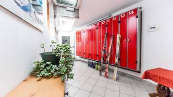 A locker room with red lockers numbered 1 to 14, a wooden bench, a table with a red tablecloth, and skis leaning against the lockers.