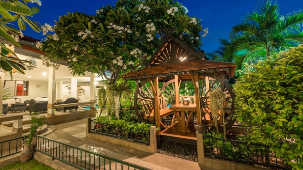 Un kiosque doté d’une structure en bois et d’une grande roue, entouré d’une végétation luxuriante et d’un espace piscine.