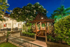 Um gazebo com estrutura de madeira e uma grande roda, rodeado por vegetação exuberante e uma área de piscina.