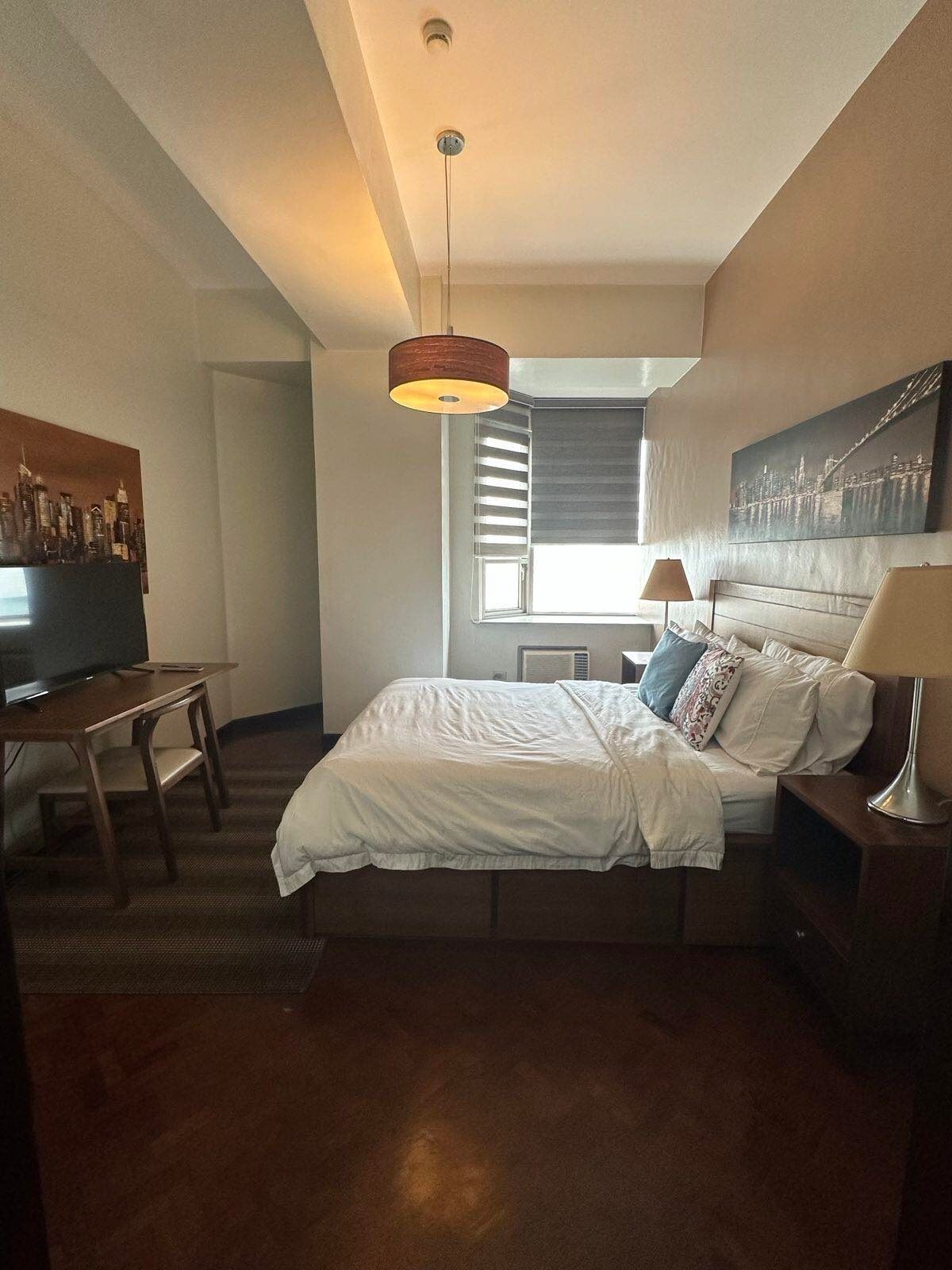 Una habitación de hotel con cama, escritorio, televisión, ventana con persianas y obras de arte en las paredes.