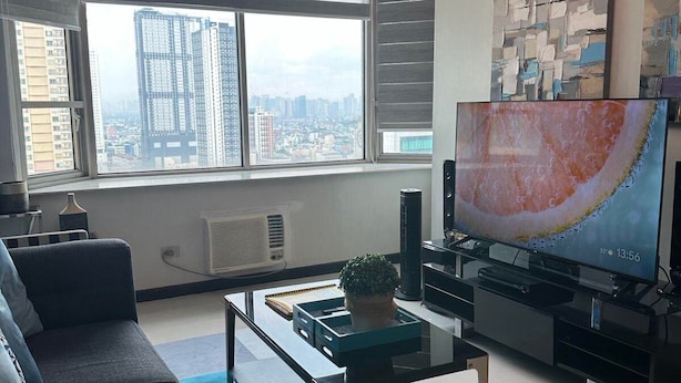 A modern living room with a large window, a sofa, a coffee table, a TV, and a chandelier. - Suites de Marina Manila (Manila)