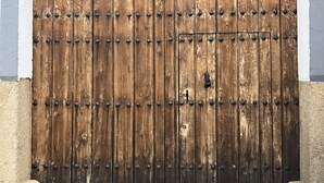 A large wooden gate with metal studs, set within a stone archway, leading to a building with a white exterior. - La Posá (La Nava de Santiago)