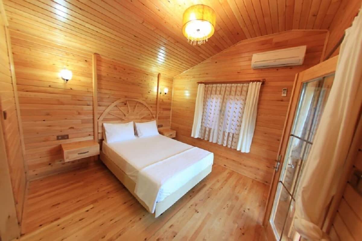 A wooden cabin bedroom with a bed, a nightstand, a wall-mounted lamp, and a window with curtains.