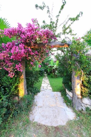 Un camino de piedra que serpentea por un jardín con un pérgola cubierta de flores rosadas.