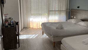 A hotel room with two beds, a wooden floor, a large window with blinds, and a small table with a kettle and a bottle. - Relais des Lilas (Parthenay)