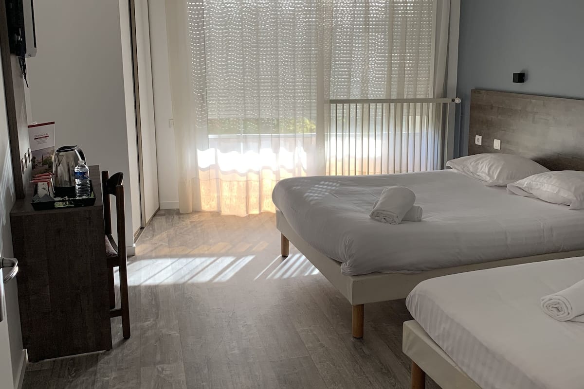 A hotel room with two beds, a wooden floor, a large window with blinds, and a small table with a kettle and a bottle.