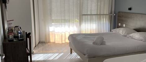 A hotel room with two beds, a wooden floor, a large window with blinds, and a small table with a kettle and a bottle.