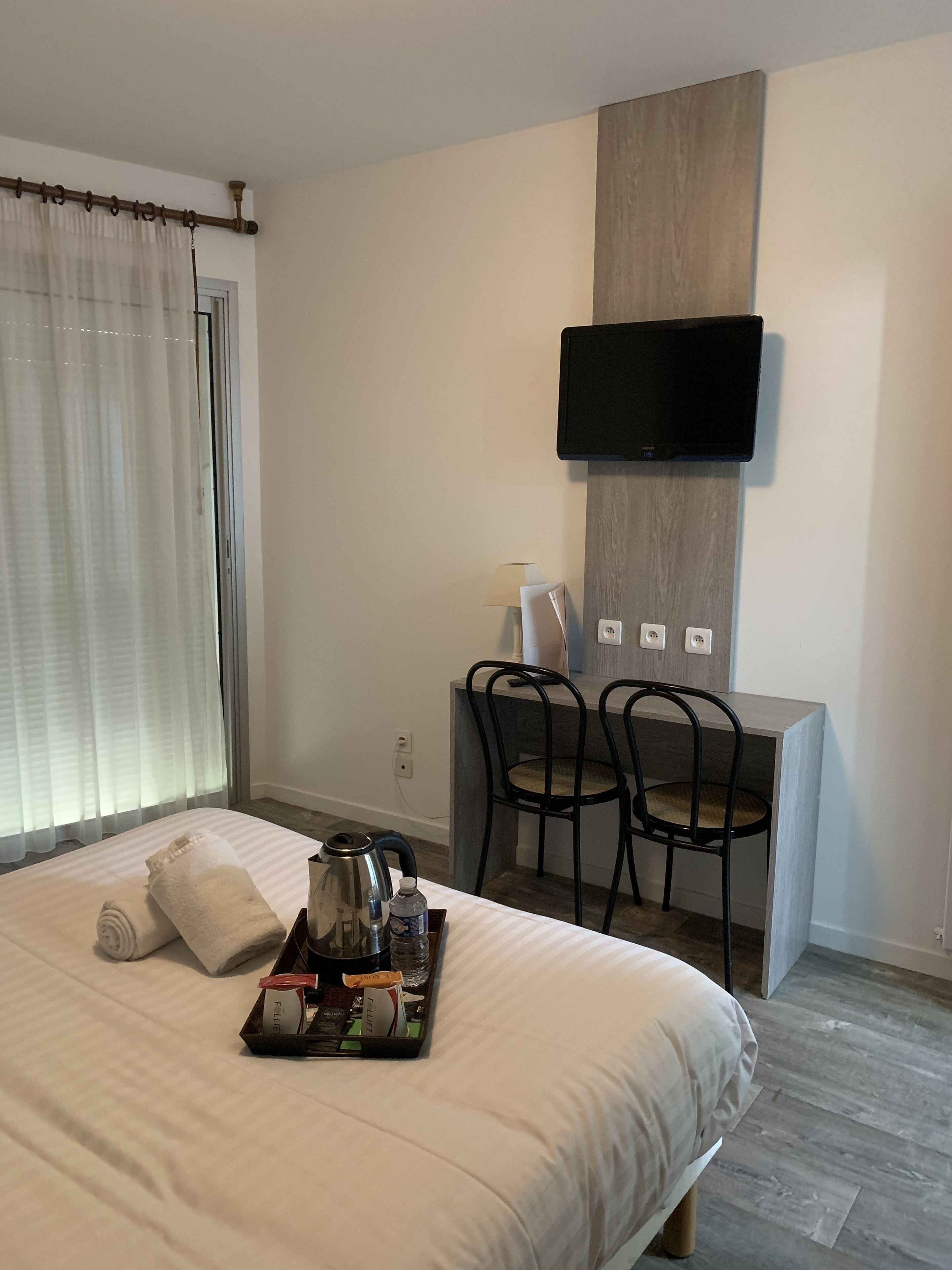 A hotel room with a bed, a desk with two chairs, a flat-screen TV mounted on the wall, and a window with curtains.
