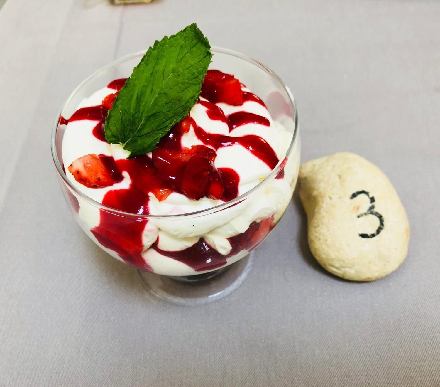 Ein Dessert in einer klaren Glaskugel mit Sahne, Erdbeeren und roter Sauce, dazu ein Keks mit der Aufschrift 3.