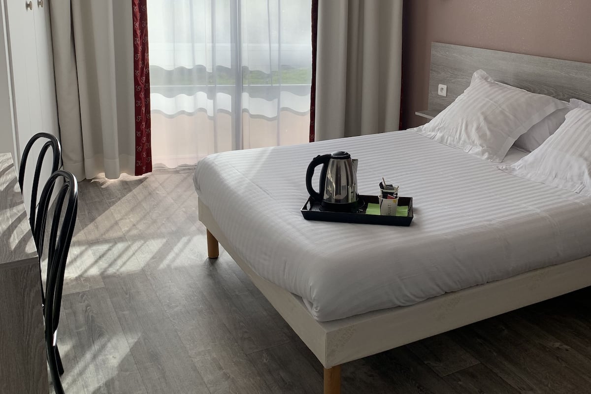 A hotel room with a large bed, a bedside table with a kettle, and a view of the outdoors through sheer curtains.