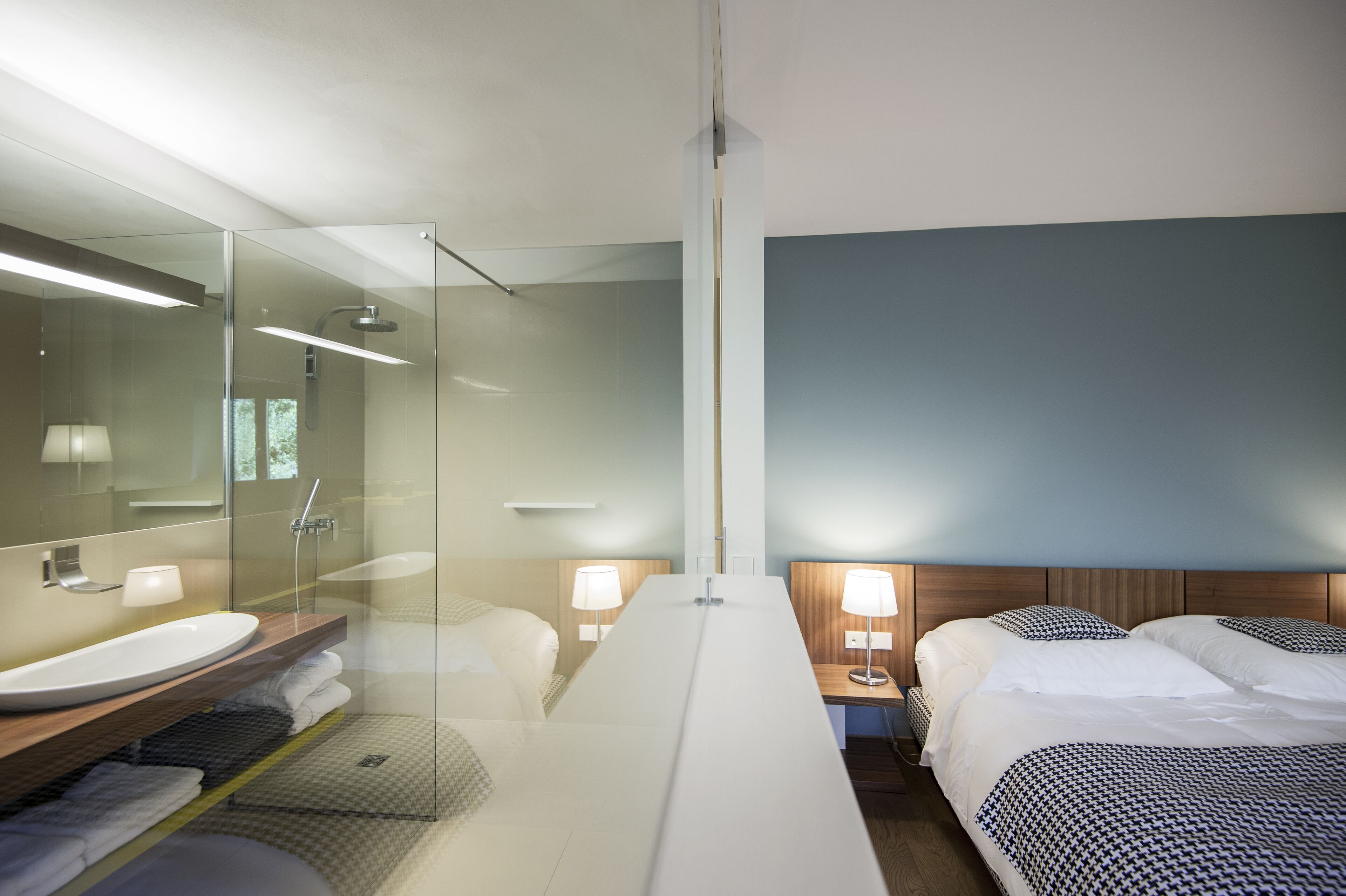 A modern hotel room with a glass shower enclosure, a large bed with patterned bedding, a wooden nightstand with a lamp, and a large mirror above the sink.