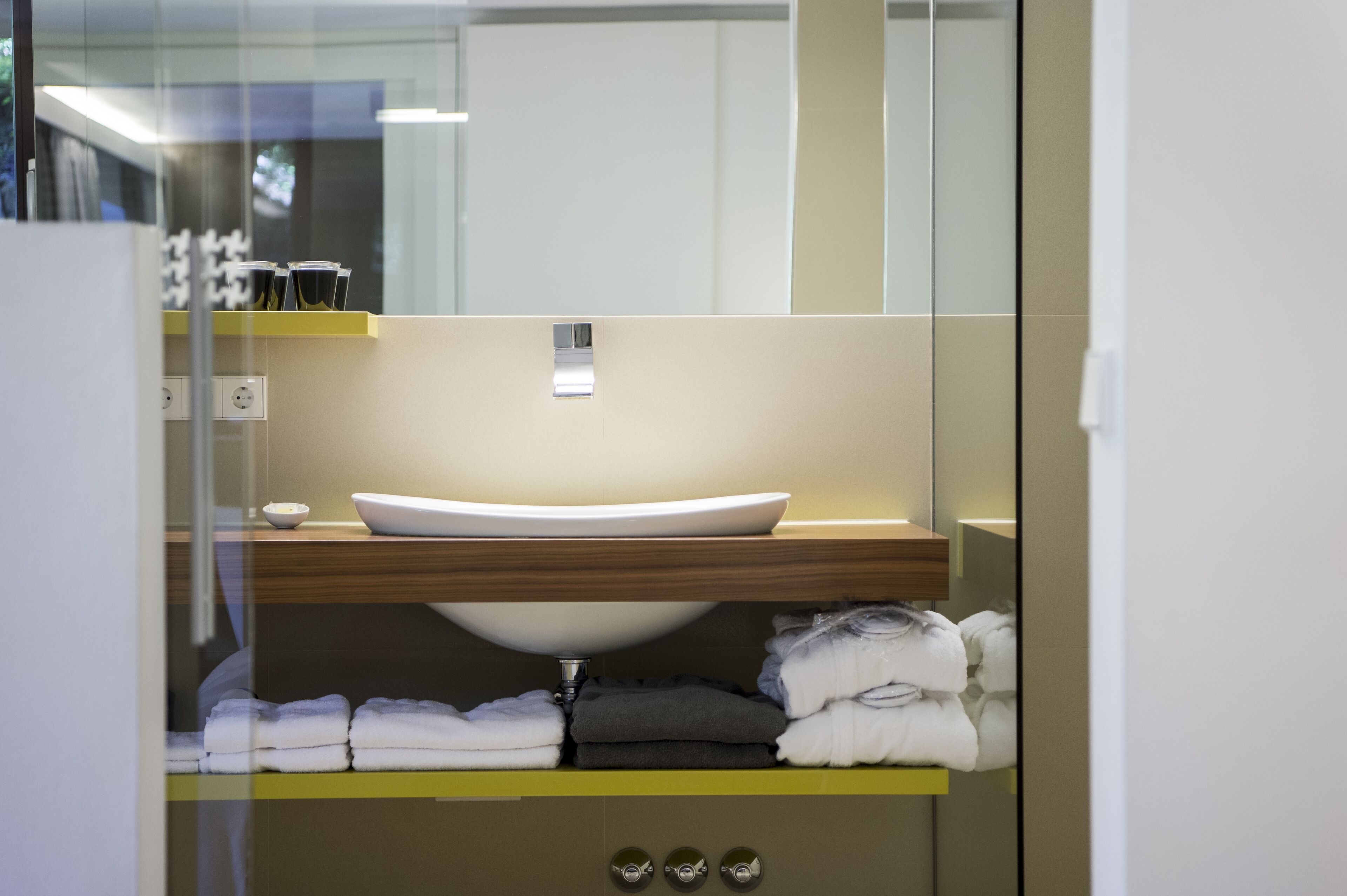 Une salle de bain moderne avec un lavabo sur colonne blanc, un miroir au-dessus et des serviettes soigneusement pliées sur une étagère en dessous.