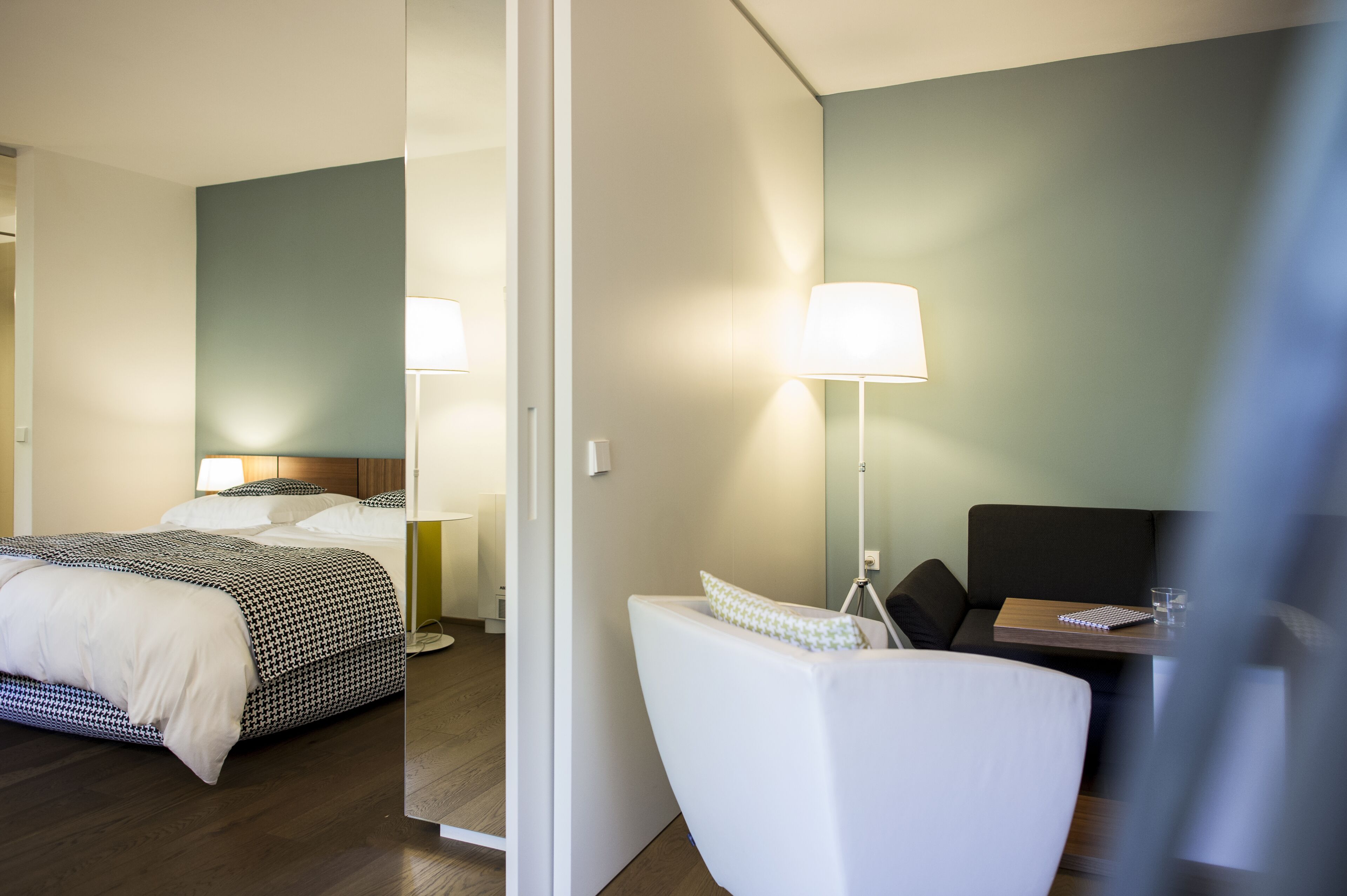 A hotel room with a bed, bedside table, lamp, and a small seating area with a chair and table.