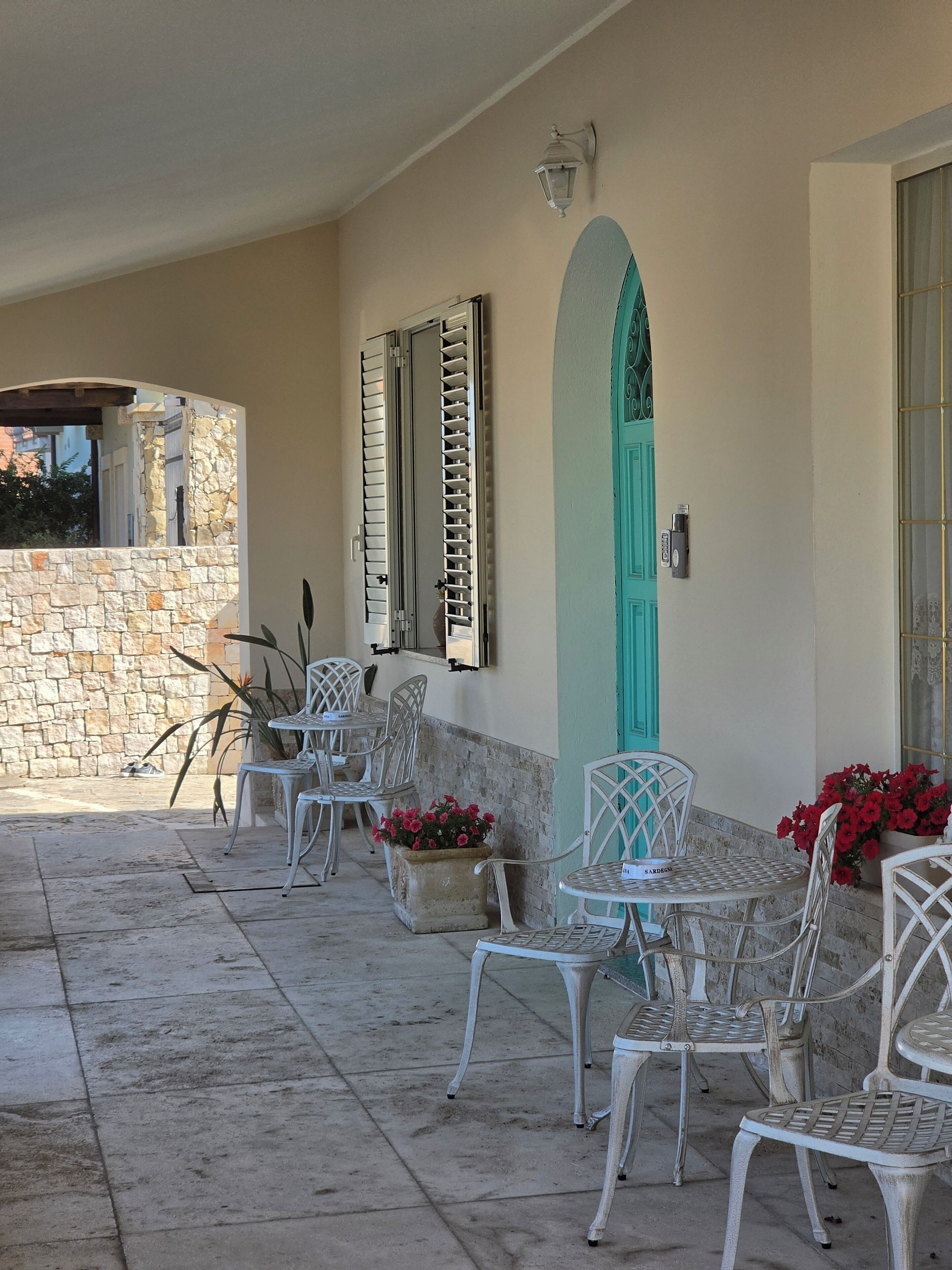 A covered outdoor seating area with stone walls, metal chairs, a turquoise door, and a stone wall with a window.