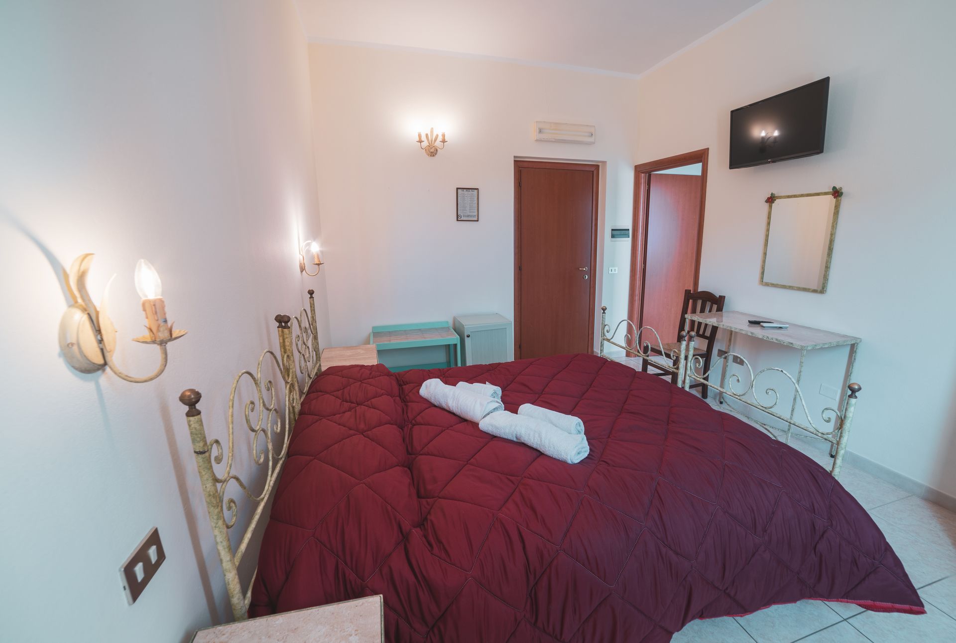 A bedroom with a large bed, a bedside table, a small table, a mirror, and a TV on the wall.