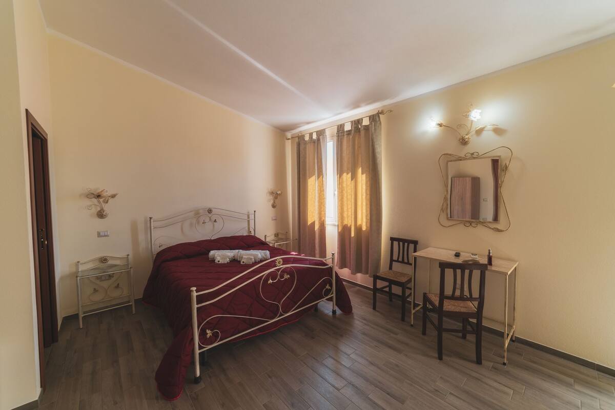Une chambre à coucher avec un lit, un bureau, une chaise, un miroir et une fenêtre avec des rideaux.