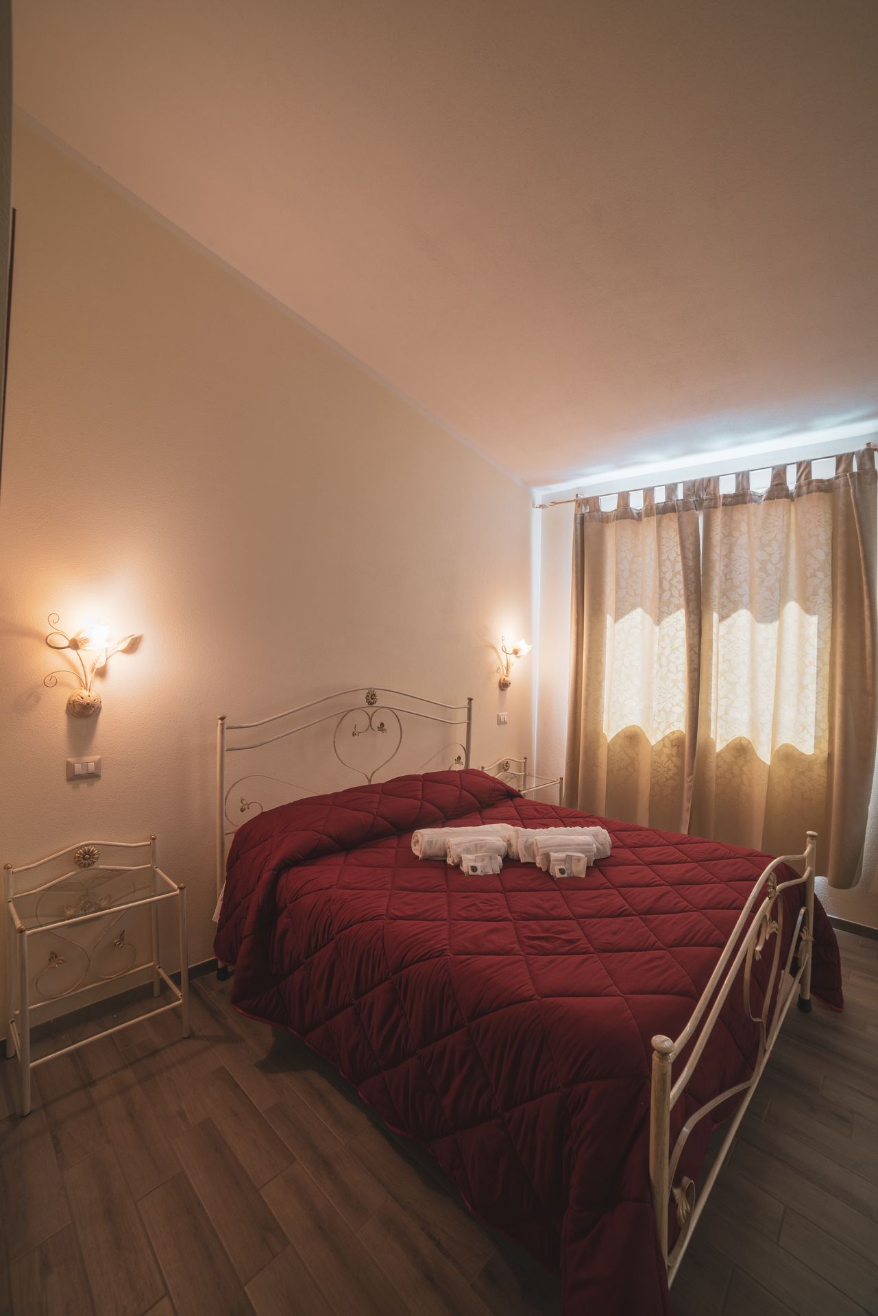 Un dormitorio con estructura metálica para la cama, una colcha acolchada roja, una mesita de noche con lámpara y una ventana con cortinas.