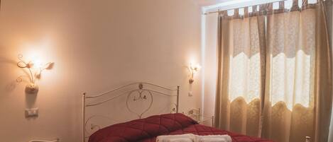 Un dormitorio con estructura metálica para la cama, una colcha acolchada roja, una mesita de noche con lámpara y una ventana con cortinas.