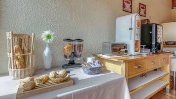 A breakfast setup with bread, pastries, and condiments on a table, a refrigerator, and a coffee maker.