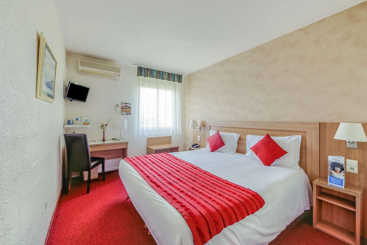 A hotel room with a large bed, a desk, a chair, a TV, a window with curtains, and a red carpet.