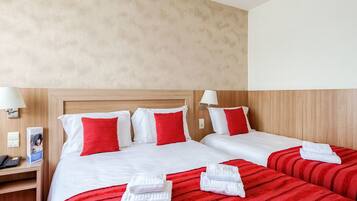 Habitación de hotel con dos camas, cada una con ropa blanca y almohadas rojas. Se observa un mesita de noche con una lámpara y un teléfono.