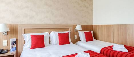 Habitación de hotel con dos camas, cada una con ropa blanca y almohadas rojas. Se observa un mesita de noche con una lámpara y un teléfono.
