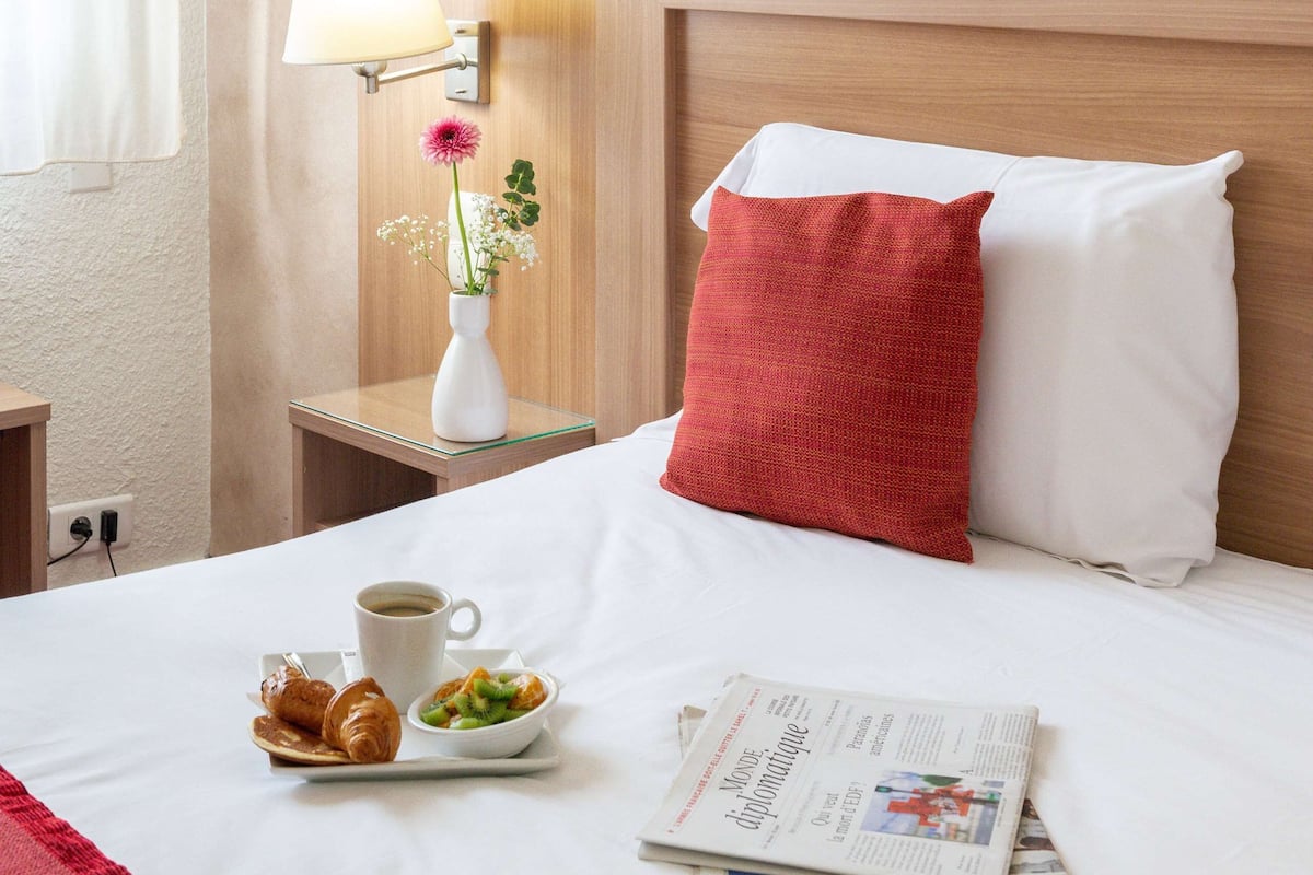 A hotel room with a neatly made bed, a bedside table with a lamp, a vase with flowers, a tray with croissants and a cup of coffee, and a newspaper.
