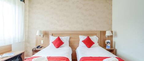 Habitación de hotel con dos camas, cada una con colcha a rayas rojas y almohadas blancas. Hay dos mesitas de noche con lámparas, un teléfono y una pequeña estantería con un libro y una revista.