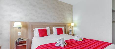 Habitación de hotel con una cama grande, ropa de cama roja y blanca, dos lámparas de noche, una mesita de noche de madera con un teléfono y una manta de rayas rojas.