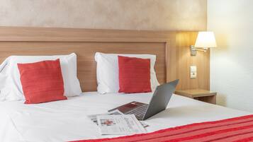 Una cama bien hecha con almohadas rojas y blancas, una computadora portátil y una revista sobre el catre de una habitación de hotel.