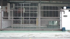 A metal gate with a curved top, a number "32" on the wall to the right, and a blue trash bin beside it. - 32 First Island (George Town)